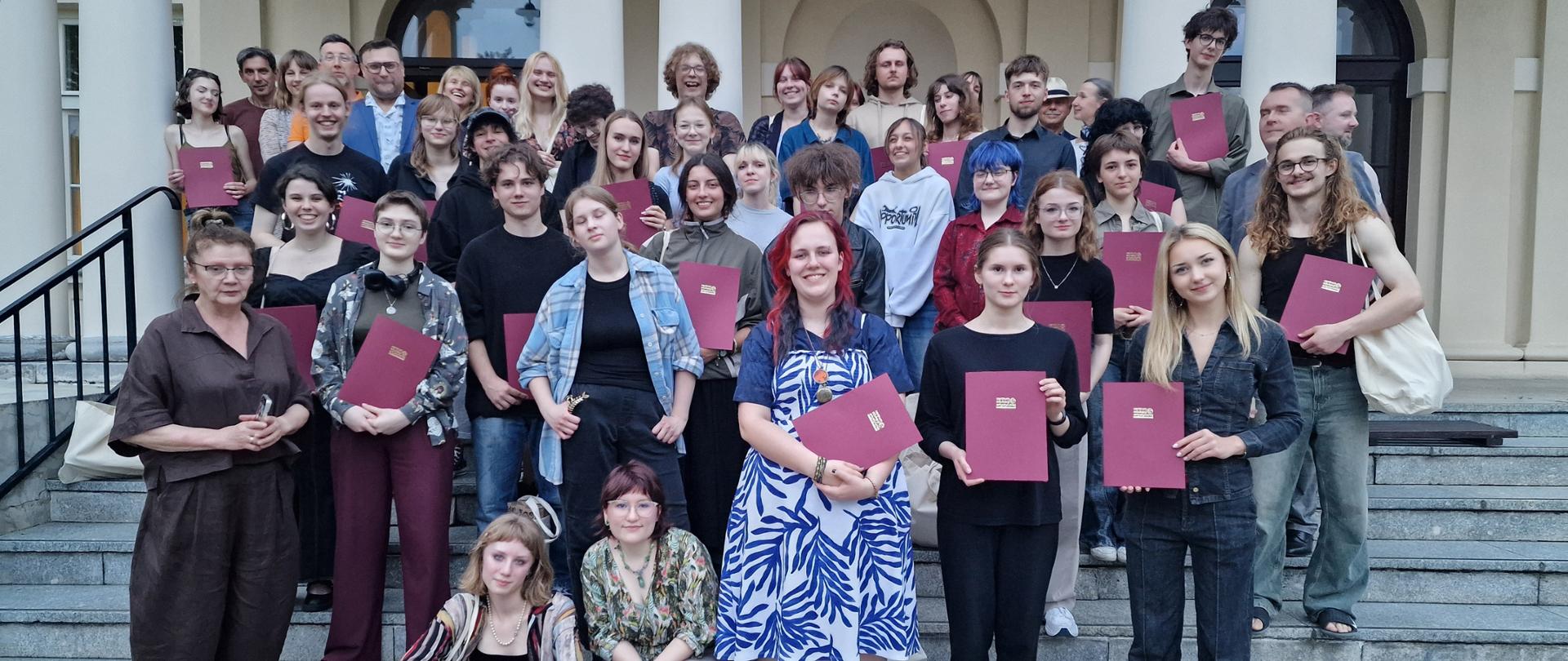 Obraz przedstawia dużą grupę młodych ludzi i kilka starszych osób stojących i siedzących na szerokich, kamiennych schodach przed dużym budynkiem. Większość z nich trzyma w rękach bordowe teczki lub certyfikaty.