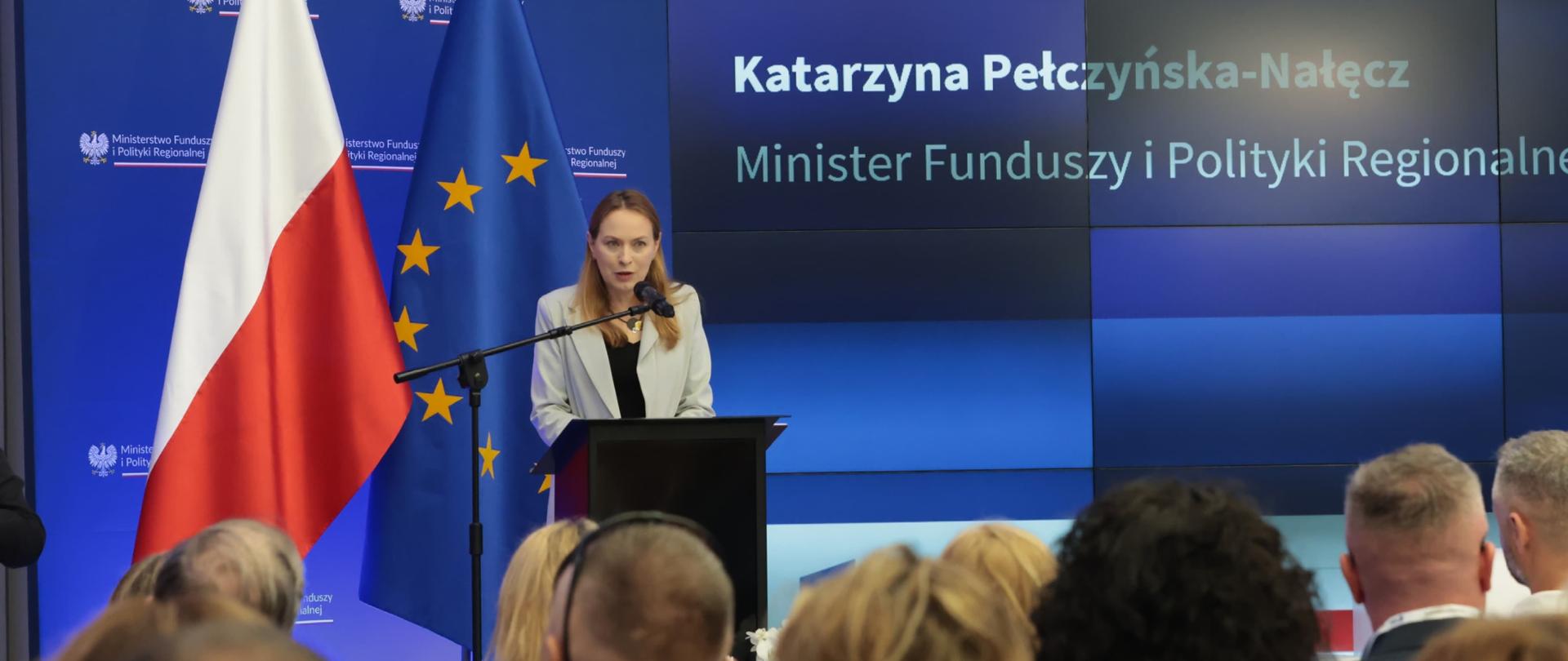 Minister Katarzyna Pełczyńska-Nałęcz w mównicy z mikrofonem na tle flag PL i UE i ścianki. Przed nią siedzi na publiczność.